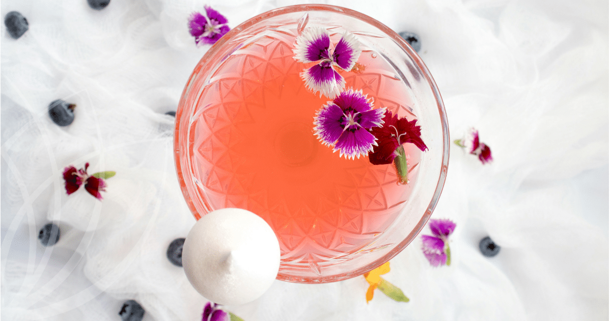 Klaasist kokteiliklaasis roosa joogi, valgete ja lillade õite ning mustikatega, taustaks valge pitsiline kangas.